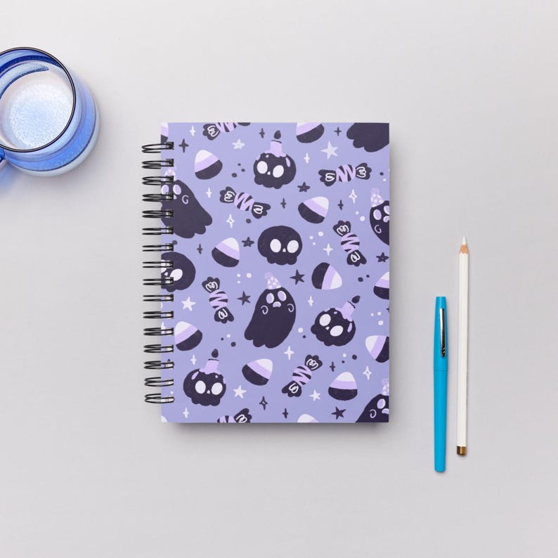 May include: Spiral-bound notebook with a lavender cover featuring a Halloween-themed pattern. The design includes black ghosts, skulls, candy corn, and wrapped candies. A blue pen and white pencil are next to the notebook.