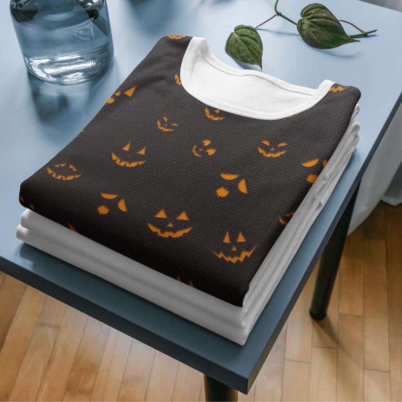May include: A stack of folded black shirts with orange jack-o'-lantern faces. The shirts have a white collar and are stacked on top of white shirts. The shirts are on a blue table.