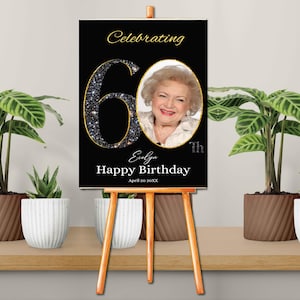 May include: Black and gold 60th birthday sign with a photo of a woman. The sign says "Celebrating 60th Evelyn Happy Birthday April 20 XXXX".