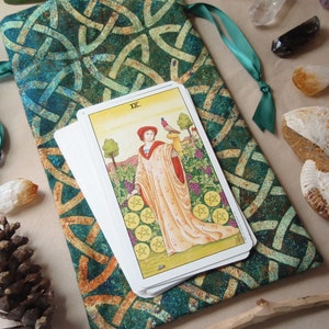 Puede incluir: Una bolsa de tela verde y dorada con un diseño de nudo celta, que contiene una baraja de cartas de tarot. La carta superior es el Nueve de Pentáculos, que muestra a una mujer en un jardín con un pájaro en la mano.