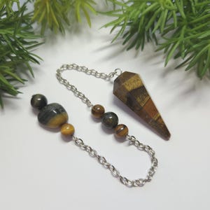 May include: A pendulum with a silver chain and a faceted, brown and tan crystal point. The chain connects to a series of small, round, and oval-shaped tiger's eye and gray-striped stones. Green foliage is in the background.