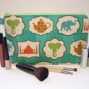 Puede incluir: Una bolsa de cremallera de color turquesa con un estampado colorido de elefantes, teteras y un palacio indio. La bolsa está rodeada de pinceles de maquillaje y tubos de maquillaje.