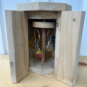 May include: A wooden key holder with a rotating spindle inside. The holder has a hinged door that opens to reveal the spindle with keys hanging from it. The holder is made of unfinished wood and has a natural wood color.