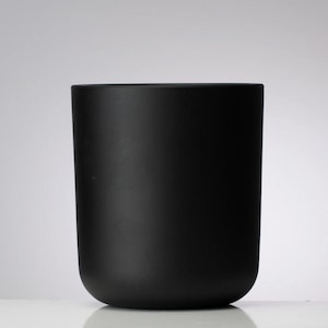 May include: A matte black cylindrical container with a rounded base. The container has a smooth, non-reflective surface and a simple, minimalist design. The object is likely a candle holder or a decorative vase.