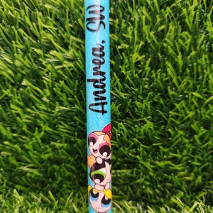 May include: A blue glitter pen featuring the text "Andrea, SW" in black script. The pen is decorated with a cartoon image of the Powerpuff Girls. The pen has a silver tip and a black base.