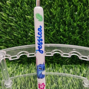 May include: A white glitter pen with the name "Jessica" in blue script. The pen features a green leaf design, a blue cartoon character, and a pink flower. The pen is displayed on a clear plastic stand against a green background.