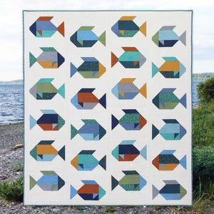 May include: A white quilt featuring a repeating pattern of colorful fish. Each fish is composed of geometric shapes in shades of blue, green, orange, and teal. The quilt has a teal border and is displayed outdoors near water.