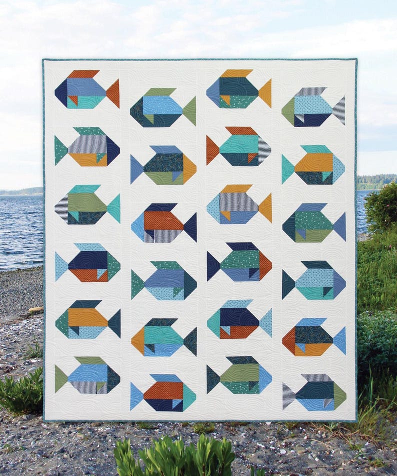 May include: A white quilt featuring a repeating pattern of colorful fish. Each fish is composed of geometric shapes in shades of blue, green, orange, and teal. The quilt has a teal border and is displayed outdoors near water.
