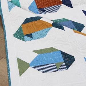 May include: A white quilt featuring a repeating pattern of stylized fish. The fish are composed of various geometric shapes in shades of blue, green, and orange, with patterned details. The quilt has a teal border.