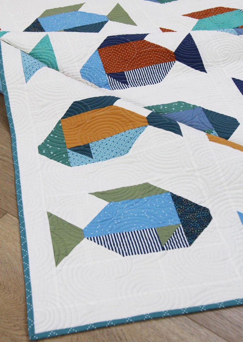May include: A white quilt featuring a repeating pattern of stylized fish. The fish are composed of various geometric shapes in shades of blue, green, and orange, with patterned details. The quilt has a teal border.