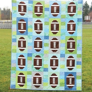 May include: A quilt pattern featuring a football design. The quilt is made up of brown footballs on a blue, green, and white background. The quilt is titled "Touchdown" and is number 143. The quilt pattern is from Cluck Cluck Sew.