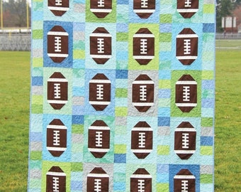 May include: A quilt pattern featuring a football design. The quilt is made up of brown footballs on a blue, green, and white background. The quilt is titled "Touchdown" and is number 143. The quilt pattern is from Cluck Cluck Sew.