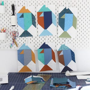 May include: Six fabric fish wall hangings with geometric designs in shades of blue, orange, and green. The fish are displayed on a white pegboard. Sewing supplies and fabric scraps are on a blue cutting mat in the foreground.
