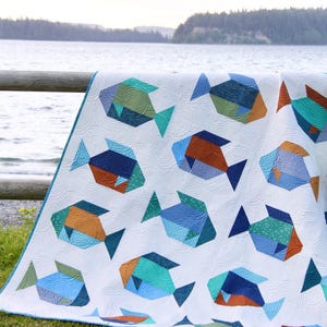 May include: A white quilt featuring a repeating pattern of geometric fish in shades of blue, teal, and orange. The quilt has a blue border and is displayed outdoors near a lake. The fish designs have textured details.