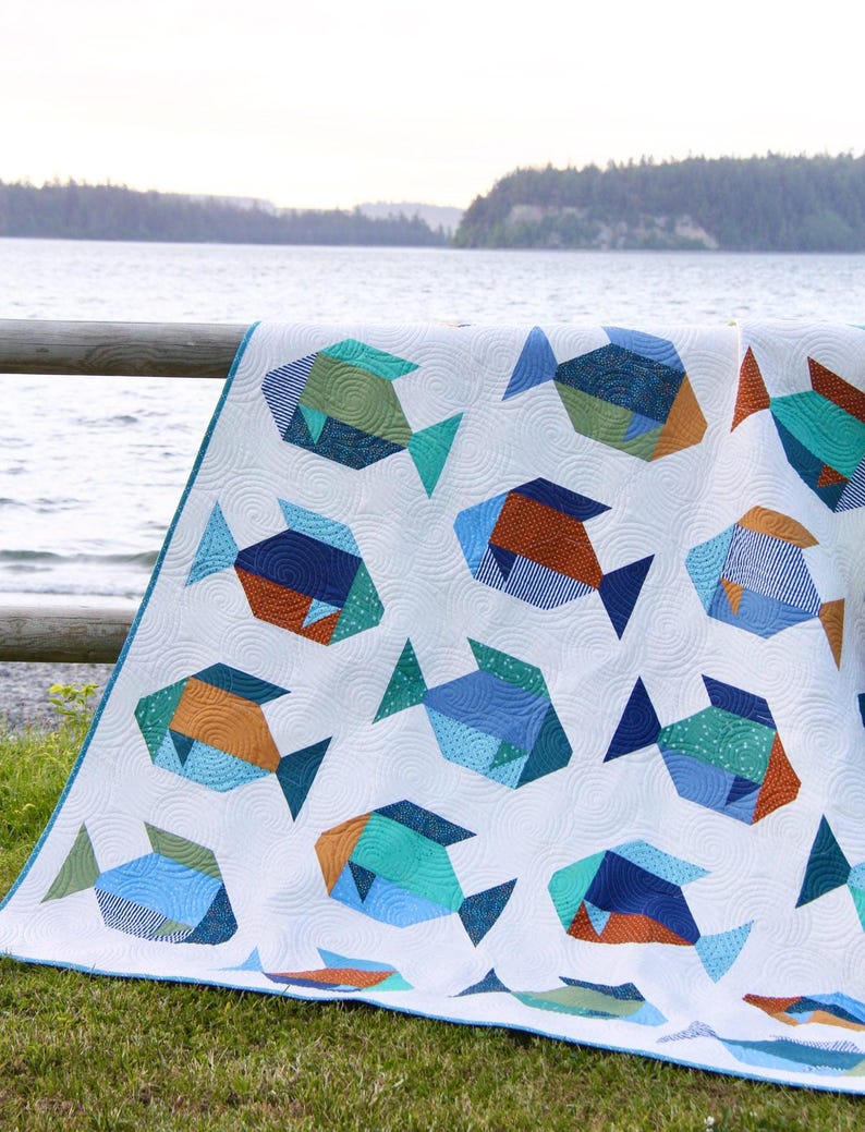 May include: A white quilt featuring a repeating pattern of geometric fish in shades of blue, teal, and orange. The quilt has a blue border and is displayed outdoors near a lake. The fish designs have textured details.