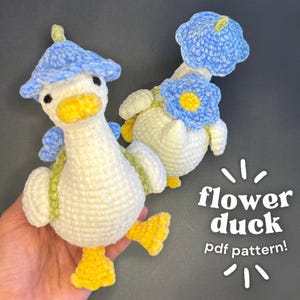 May include: Two handmade crochet ducks. One duck is upright, the other is lying down. Both are white with yellow beaks and feet. The upright duck wears a blue flower hat and has blue wings. The lying down duck has a blue flower on its head. Text reads "flower duck pdf pattern!"