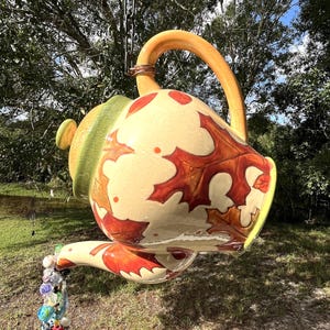 Puede incluir: Una gran tetera de cerámica colgante con un cuerpo color crema y un diseño de hojas de otoño en tonos rojos y marrones. La tetera tiene un asa y un pico amarillos, y una tapa verde. Una cadena suspende la tetera, y cuentas coloridas fluyen del pico.