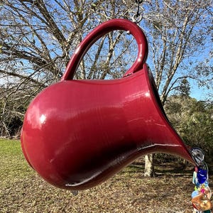 May include: A large, red, ceramic pitcher hangs upside down from a chain. The pitcher is shaped like a traditional teapot with a wide spout and a handle. The spout is pouring out a string of colorful glass beads.