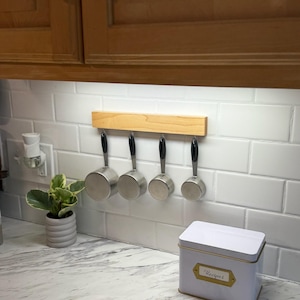May include: A kitchen scene featuring a wooden rack with four measuring cups hanging below a cabinet. A white recipe tin with a gold label that reads "Recipes" sits on the countertop. A small potted plant is also visible.