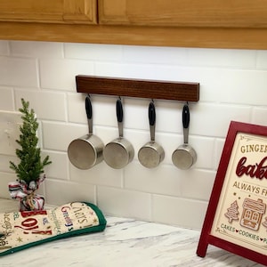 May include: A dark wood magnetic rack mounted on a white subway tile wall, holding four stainless steel measuring cups with black handles. The cups are arranged in a row, hanging from the magnetic strip by their metal handles.
