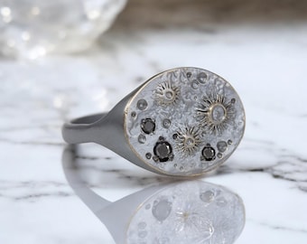May include: A silver signet ring with a textured oval top featuring a design of black diamonds and gold accents, resembling a celestial scene.
