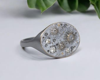 May include: A silver oval ring with a textured surface and small white diamonds set in the metal. The ring has a unique design that resembles a celestial body.