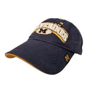 Puede incluir: Una gorra de béisbol azul marino con el texto bordado MICHIGAN WOLVERINES en letras blancas y doradas. El frente muestra un parche ovalado dorado con una letra M azul marino. Un pequeño parche cuadrado con una M dorada está en el borde.
