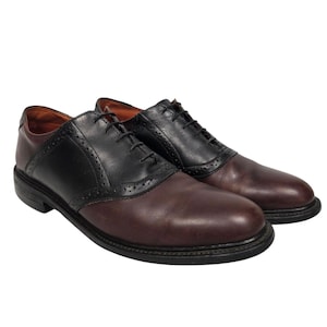 May include: A pair of two-tone oxford shoes featuring a dark brown leather toe and heel counter contrasted with a black leather saddle and lace-up panel. The shoes include decorative brogue perforations along the saddle edges and black laces.