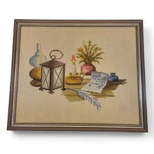 May include: A framed needlepoint embroidery depicting a still life scene with a lantern, a lit candle in a gold holder, a potted plant, a glass bottle, a small blue inkwell, a piece of parchment paper with illegible text, and a grey quill pen.