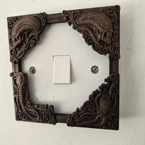 May include: A decorative brown light switch cover with ornate, gothic-style corner designs. The corners feature detailed, textured carvings of skulls and tentacles. The cover surrounds a white light switch on a white wall.