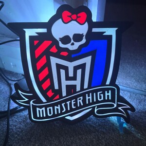 May include: A black and white Monster High logo with a red bow, a skull, and the text "Monster High" on a banner. The logo has red and blue accents and is likely a decorative item.