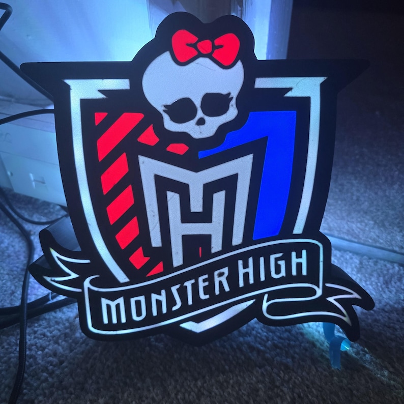 May include: A black and white Monster High logo with a red bow, a skull, and the text "Monster High" on a banner. The logo has red and blue accents and is likely a decorative item.