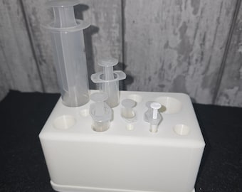 Può includere: Un organizer in plastica bianca con diverse siringhe di plastica trasparente di varie dimensioni. Le siringhe sono in posizione verticale, con pistoni visibili. L'organizer ha diversi fori per contenere le siringhe. Lo sfondo è un pannello di legno grigio chiaro.