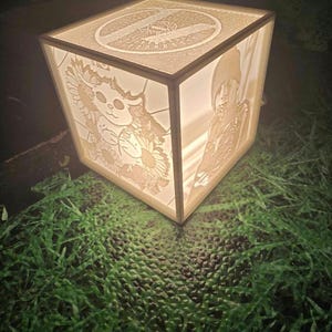 Op de afbeelding: Een witte, kubusvormige lamp met een 3D-ontwerp aan elke kant. De ontwerpen omvatten een kat met zonnebloemen, een persoon en een cirkelvormig ontwerp. De lamp is aan en geeft een warm licht.
