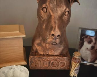 Könnte beinhalten: Eine bronzefarbene Hundebüsten-Skulptur mit einem rechteckigen Sockel, auf dessen Vorderseite der Name HUGO eingeprägt ist. Die Skulptur zeigt einen Hund mit spitzen Ohren und einer detaillierten Schnauze, neben einer Holzbox und einem Glas.