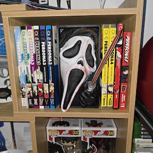 Puede incluir: Una estantería de madera exhibe una colección de manga, incluyendo volúmenes de "Persona 3" y "Persona 5". Una máscara de Scream y una espada decorativa se colocan entre los libros. Debajo, se ven dos figuras de Funko Pop.