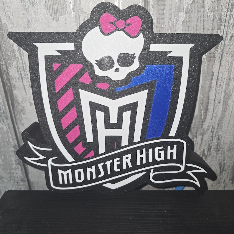 May include: A black and white Monster High logo with a pink bow, skull, and shield design. The shield has pink and blue accents, and the words "MONSTER HIGH" are displayed on a banner.