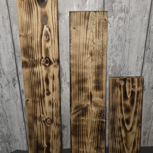 Puede incluir: Tres tablones de madera rústicos con un acabado carbonizado y texturizado. Los tablones varían en tamaño, mostrando la veta natural de la madera y los nudos. La madera tiene un color marrón oscuro con reflejos marrones claros.