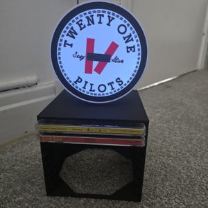 Puede incluir: Un expositor cuadrado negro con un logotipo circular de Twenty One Pilots. El logotipo presenta texto blanco, detalles en rojo y las palabras 'Stay Alive'. Dos CD están apilados en el expositor.