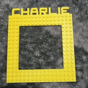 May include: A bright yellow picture frame with the name "CHARLIE" across the top. The frame is constructed from interlocking brick-style blocks, with a square opening in the center.