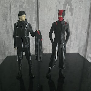 May include: Two black action figures with red accents. One figure wears a black jacket with white stripes and holds a black cape with red details. The other figure is in a black suit with a red mask and accents.