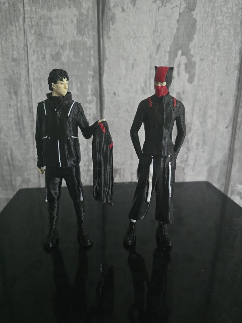 May include: Two black action figures with red accents. One figure wears a black jacket with white stripes and holds a black cape with red details. The other figure is in a black suit with a red mask and accents.