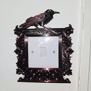 May include: A dark red, ornate light switch cover with a raven perched on top. The frame surrounds a white switch plate with a single toggle switch. The design has a gothic or vintage aesthetic, adding a decorative touch to any room.