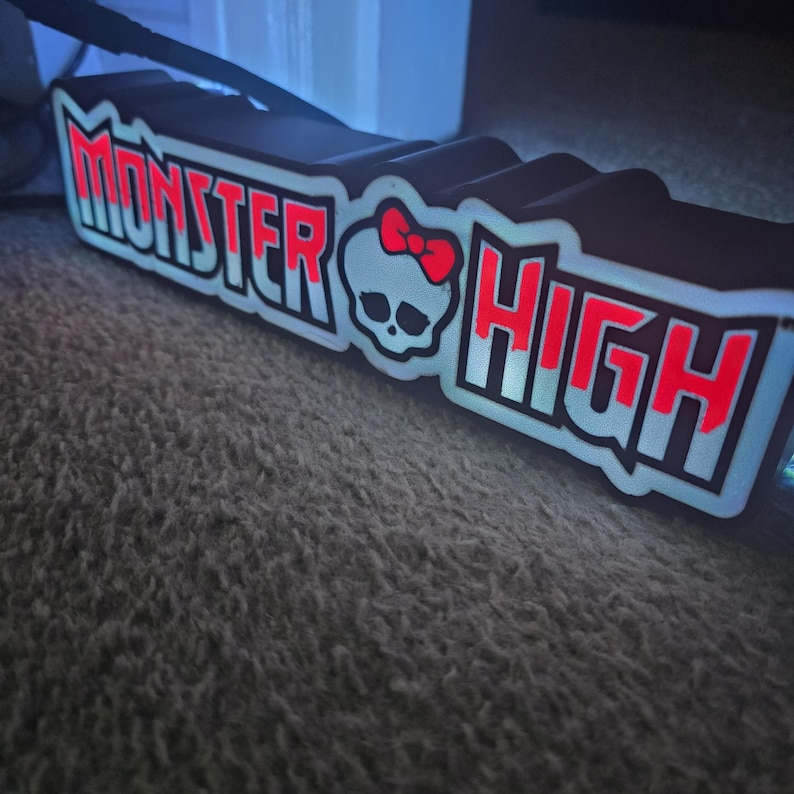 May include: A black and white Monster High sign with red lettering and a skull graphic with a red bow. The sign is illuminated with a soft glow, creating a vibrant effect. The sign is placed on a textured surface.