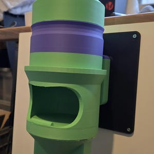 May include: A 3D-printed bird feeder, mounted on a wall, in green and purple. The feeder has a cylindrical body with a rectangular opening and a textured base. It is attached to a black mounting plate.