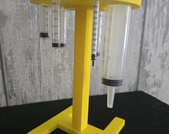 Può includere: Un supporto in plastica giallo brillante con un montante verticale centrale e una piastra superiore circolare dotata di fori multipli. Diverse siringhe in plastica trasparente di varie dimensioni sono inserite nei fori, appese verticalmente con segni di misurazione neri.