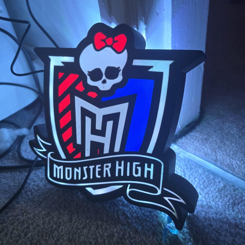 May include: A black and white Monster High logo with a skull wearing a red bow. The logo has a red and blue design with the words "Monster High" in a white banner. The logo is on a gray surface.