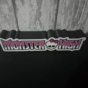 May include: A black, rectangular Monster High display stand. The text "Monster High" is in white with a pink outline, featuring a skull with a pink bow. The stand is made of a black material.