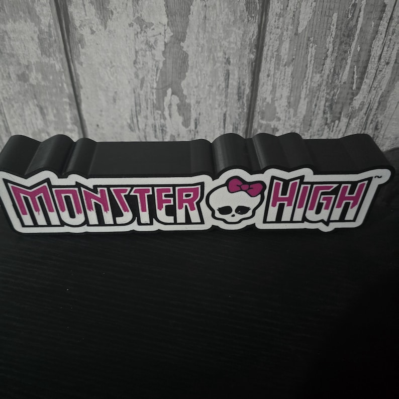 May include: A black, rectangular Monster High display stand. The text "Monster High" is in white with a pink outline, featuring a skull with a pink bow. The stand is made of a black material.