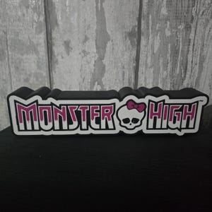 May include: A black, rectangular Monster High logo with white and pink lettering. The logo features a skull with a pink bow. The background is a light gray wooden panel.